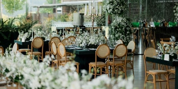 A formal event setting created by a student studying a wedding planner course with dark green tables adorned with white floral arrangements and candelabras, surrounded by natural wood chairs with woven seats.